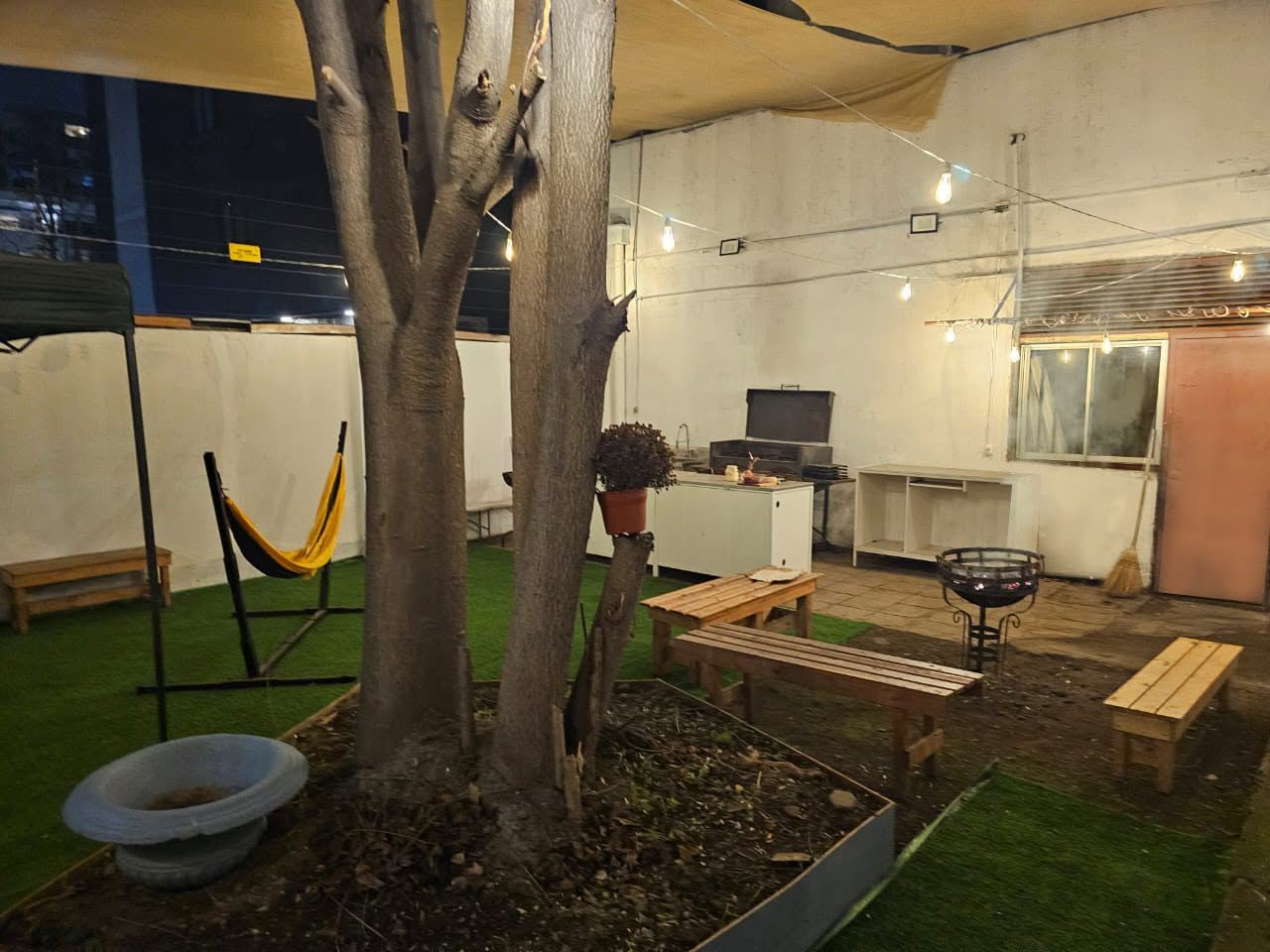 Patio exterior de KZUP de noche con parrilla, bancas de madera, hamaca y luces colgantes